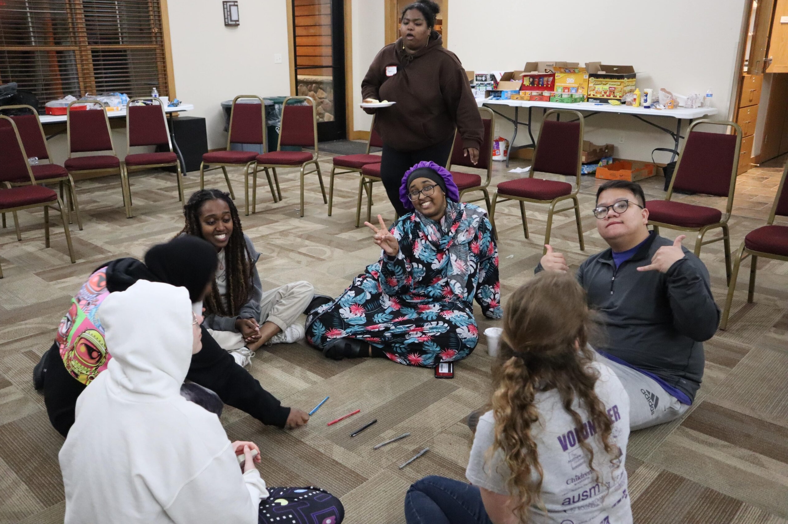 Group of students sitting on the floor in a circle with pens in the middle of them. Chairs, and a table of snacks in the background.