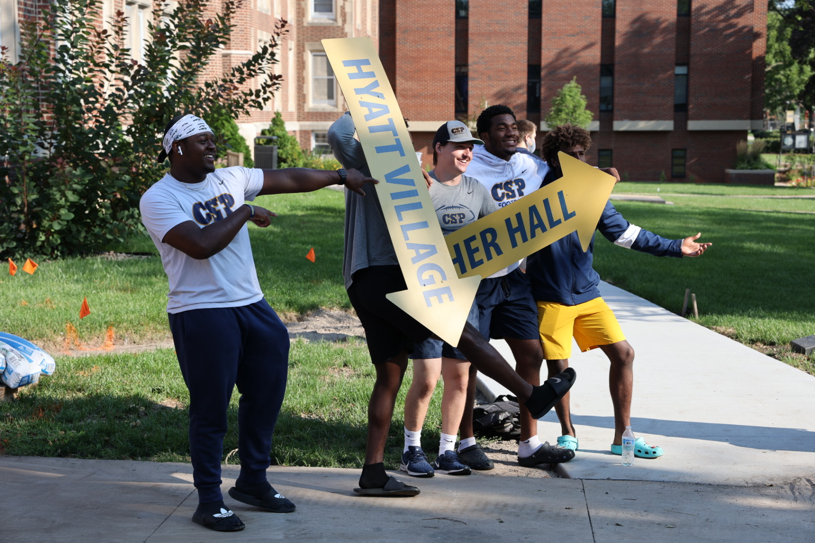 Group of college students standing on a sidewalk using arrow signs to direct people.