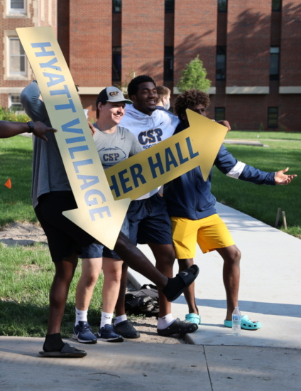 Group of college students standing on a sidewalk using arrow signs to direct people.