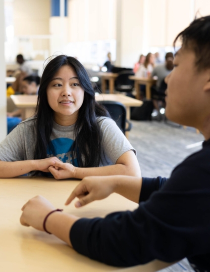 2 students sitting at a table and talking to each other.