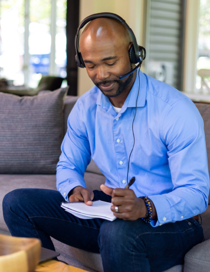 Online student sitting on couch with laptop in front of him taking notes on a notepad and wearing a headset.