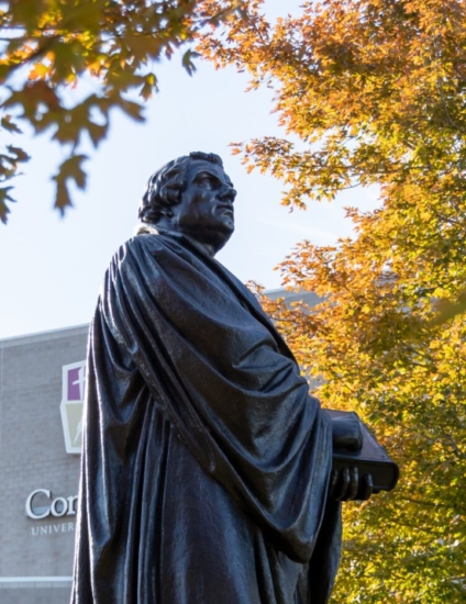 Dark bronze statue in front of an academic building with brightly colored yellow and green trees on either side of the statue.