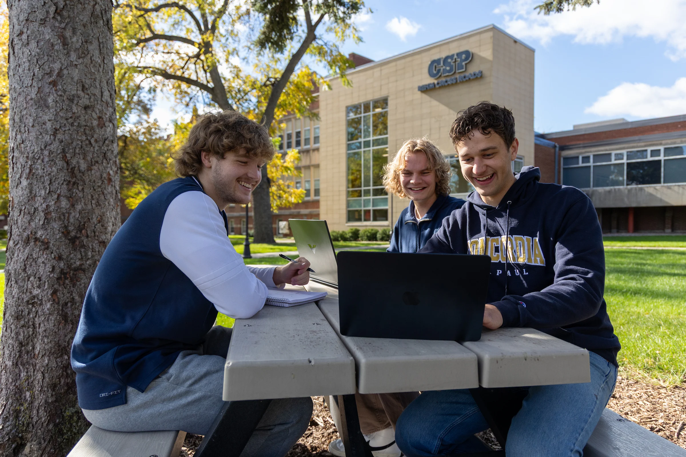 Group of 3 students sitting at a picnic table outside on campus in autumn.