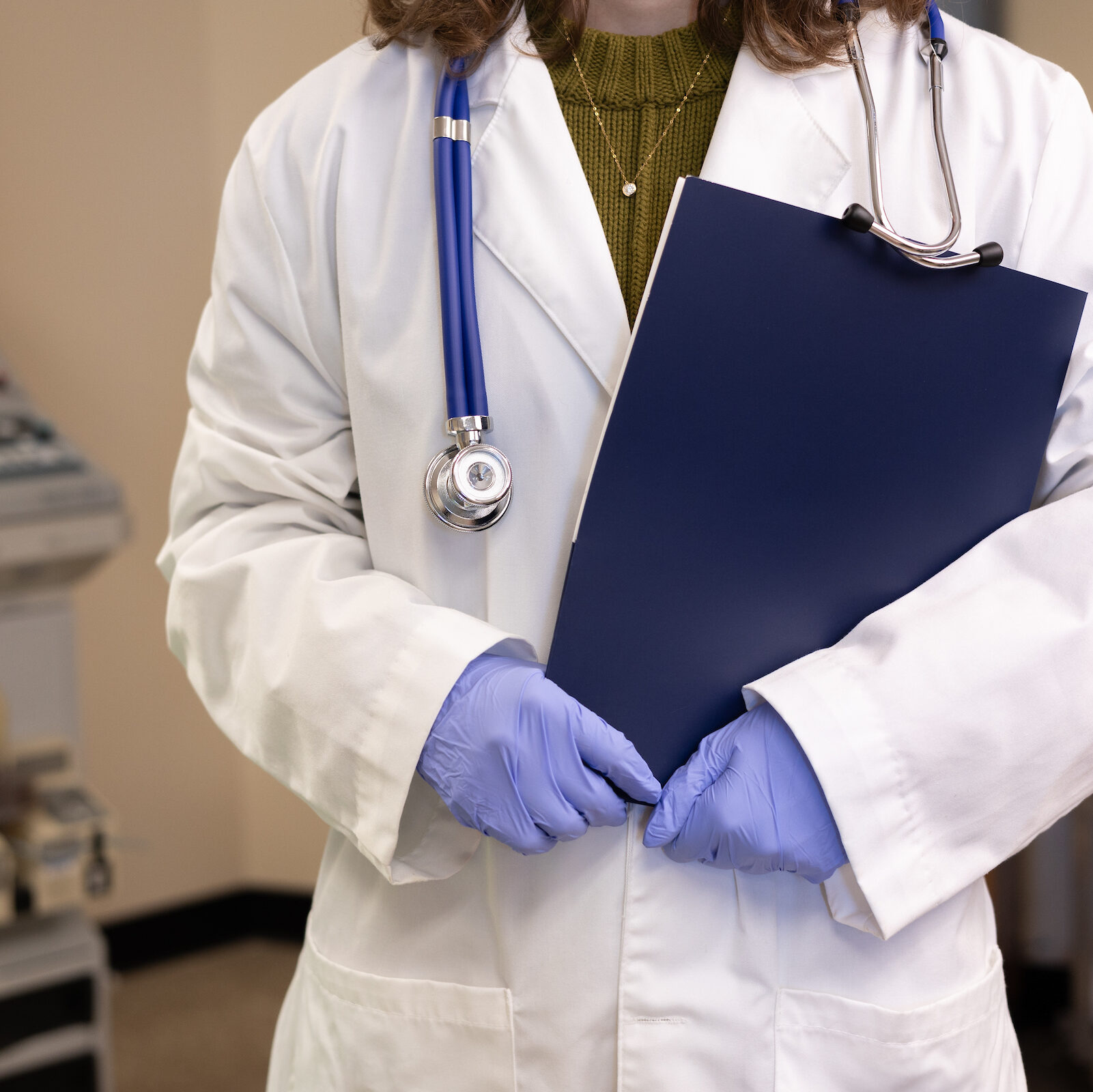 Medical professional wearing purple gloves, a white coat, and a stethoscope holding a blue folder.