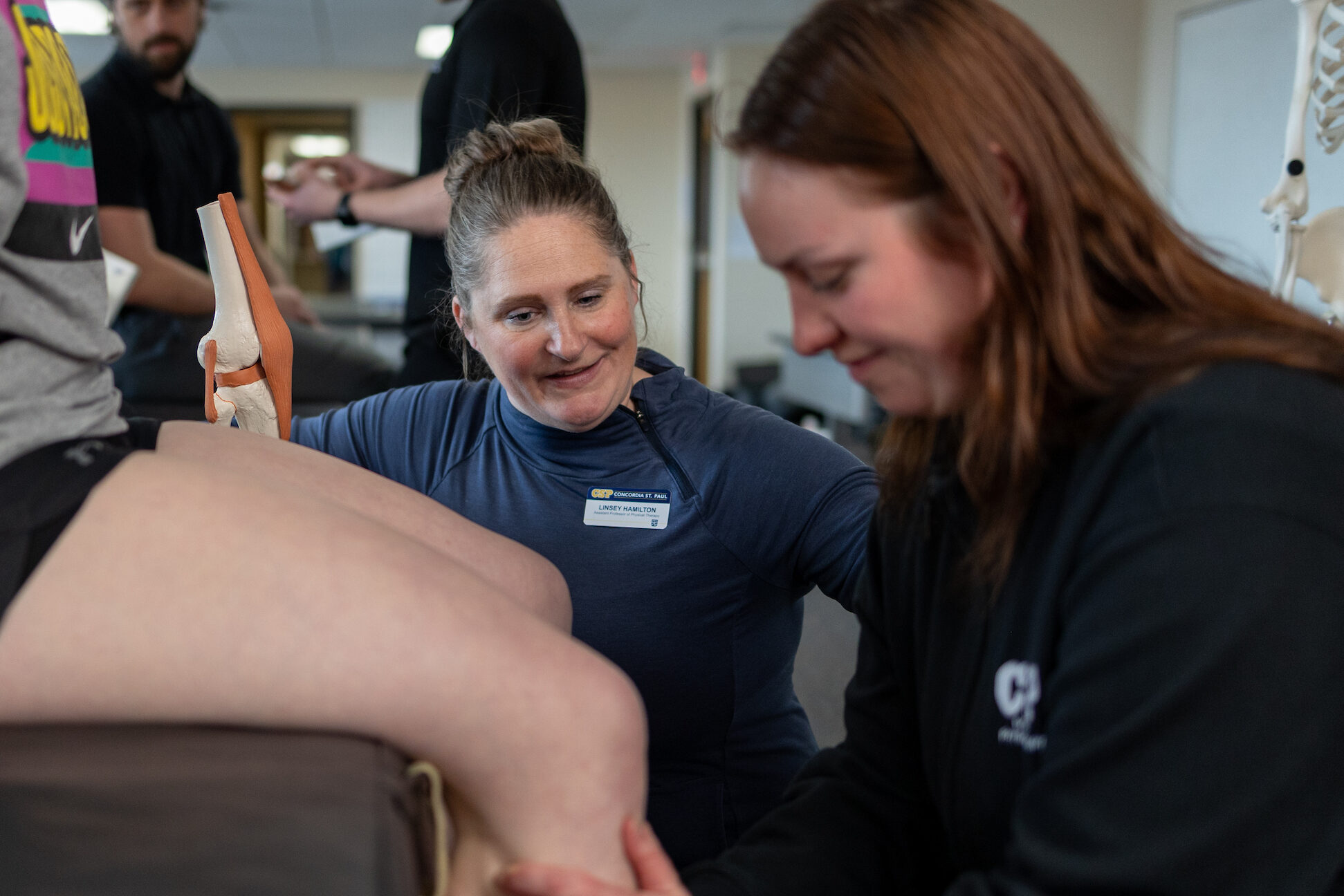 Professor and student examining a volunteer's knee in a classroom lab setting.