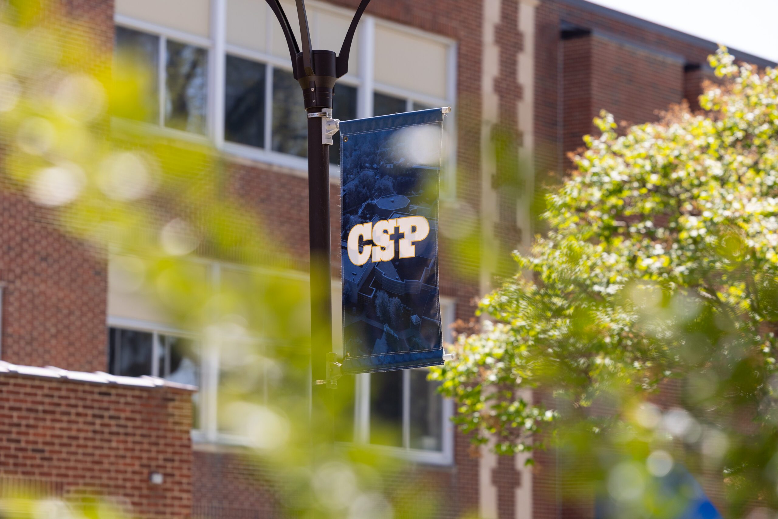 Blue CSP Banner surrounded by trees in front of a brick building.