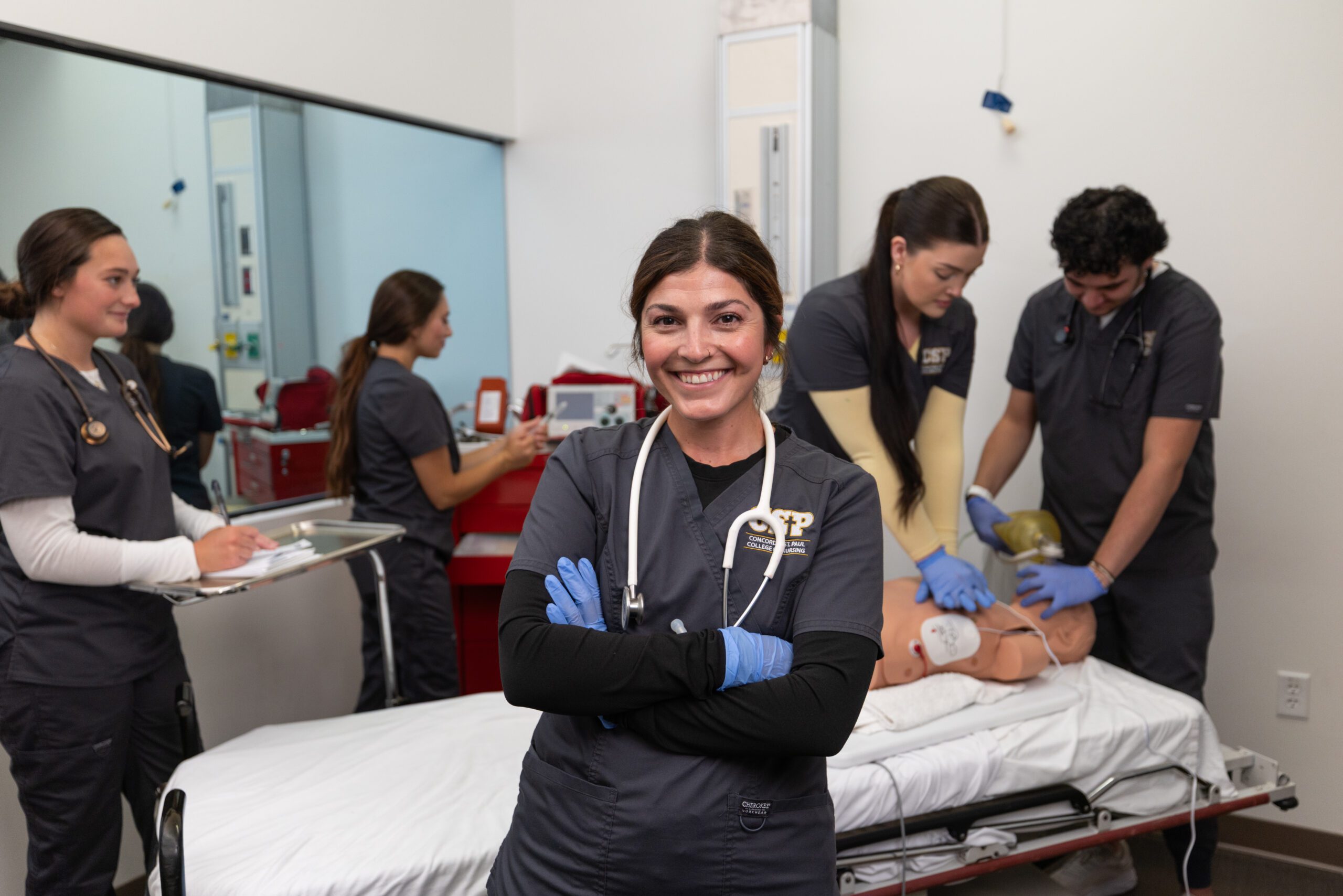 Nursing student standing with arms crossed in nursing lab with other students in the background.