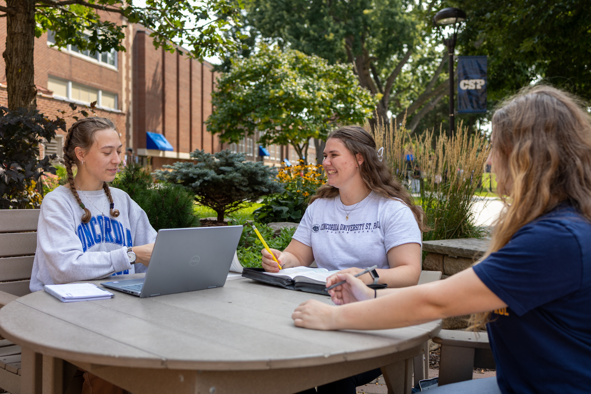 3 students sitting outside at a table using a laptop and studying books.