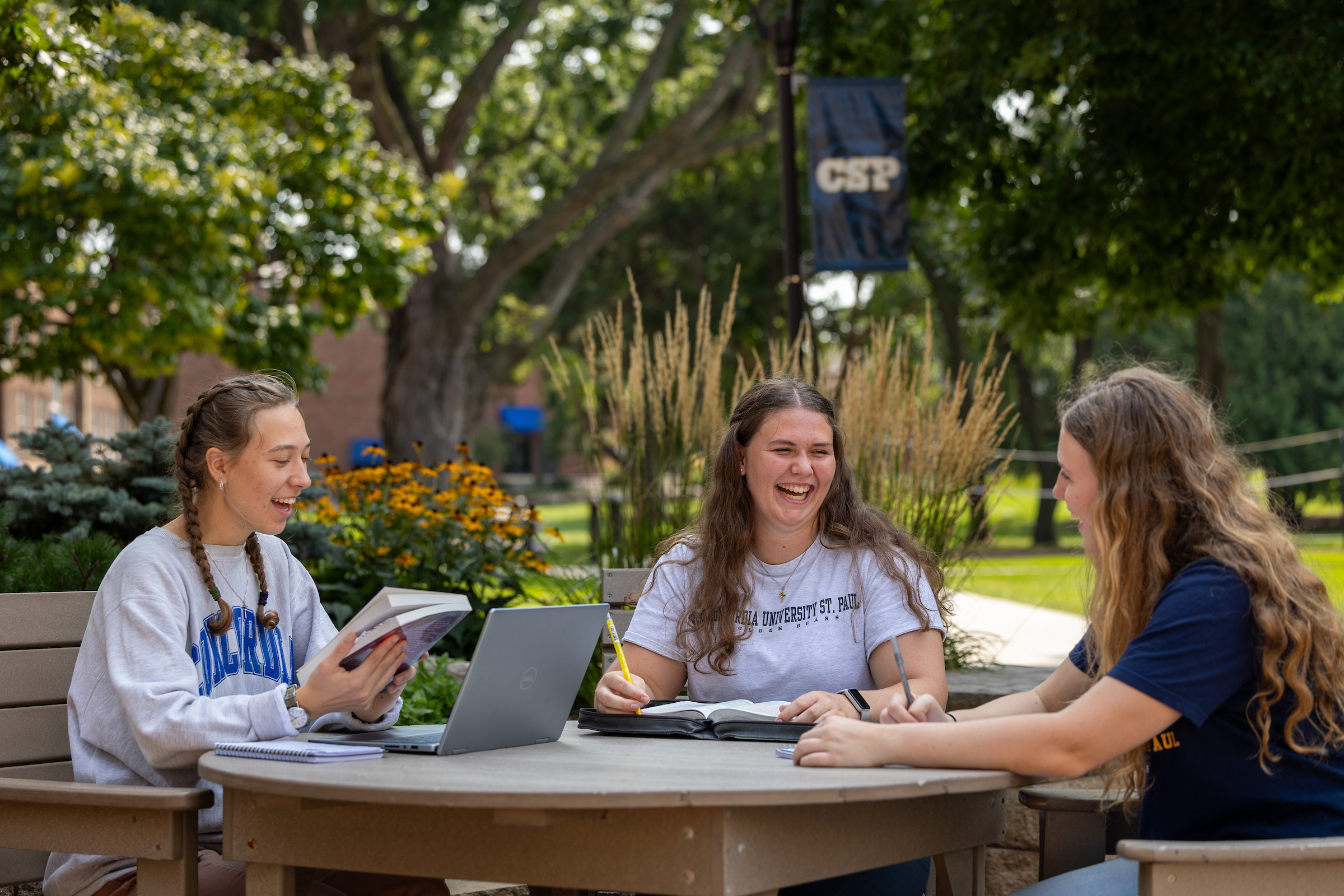3 college students sitting at a table outdoors in summer time studying books and working on a laptop.