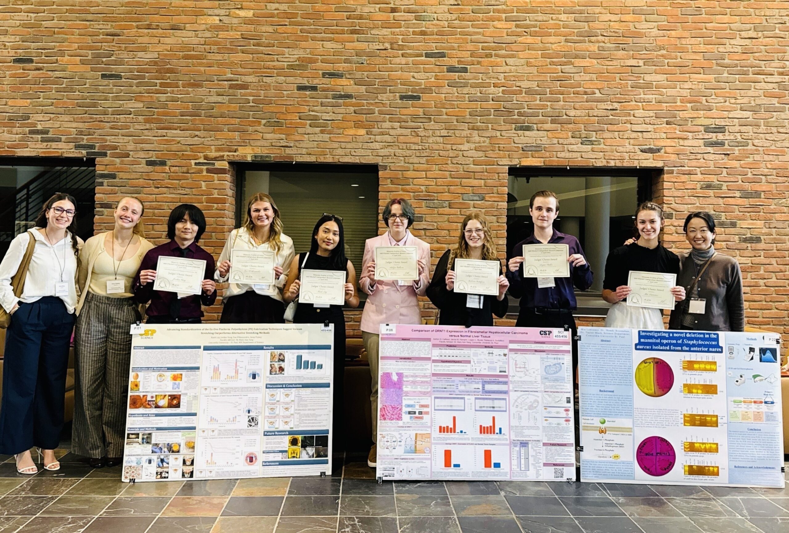 A group of CSP science students stand in front of a brick wall holding their awards and posters.