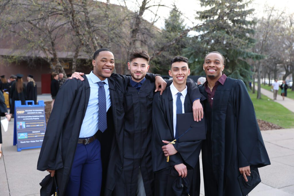 Group of four CSP students at graduation wearing commencement gowns outdoors.